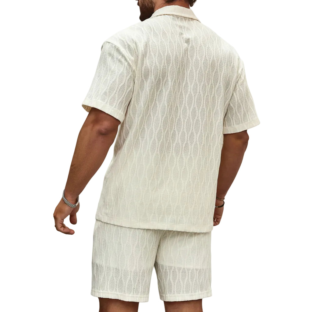 Man wearing a beige patterned shirt and shorts set against a stone wall.